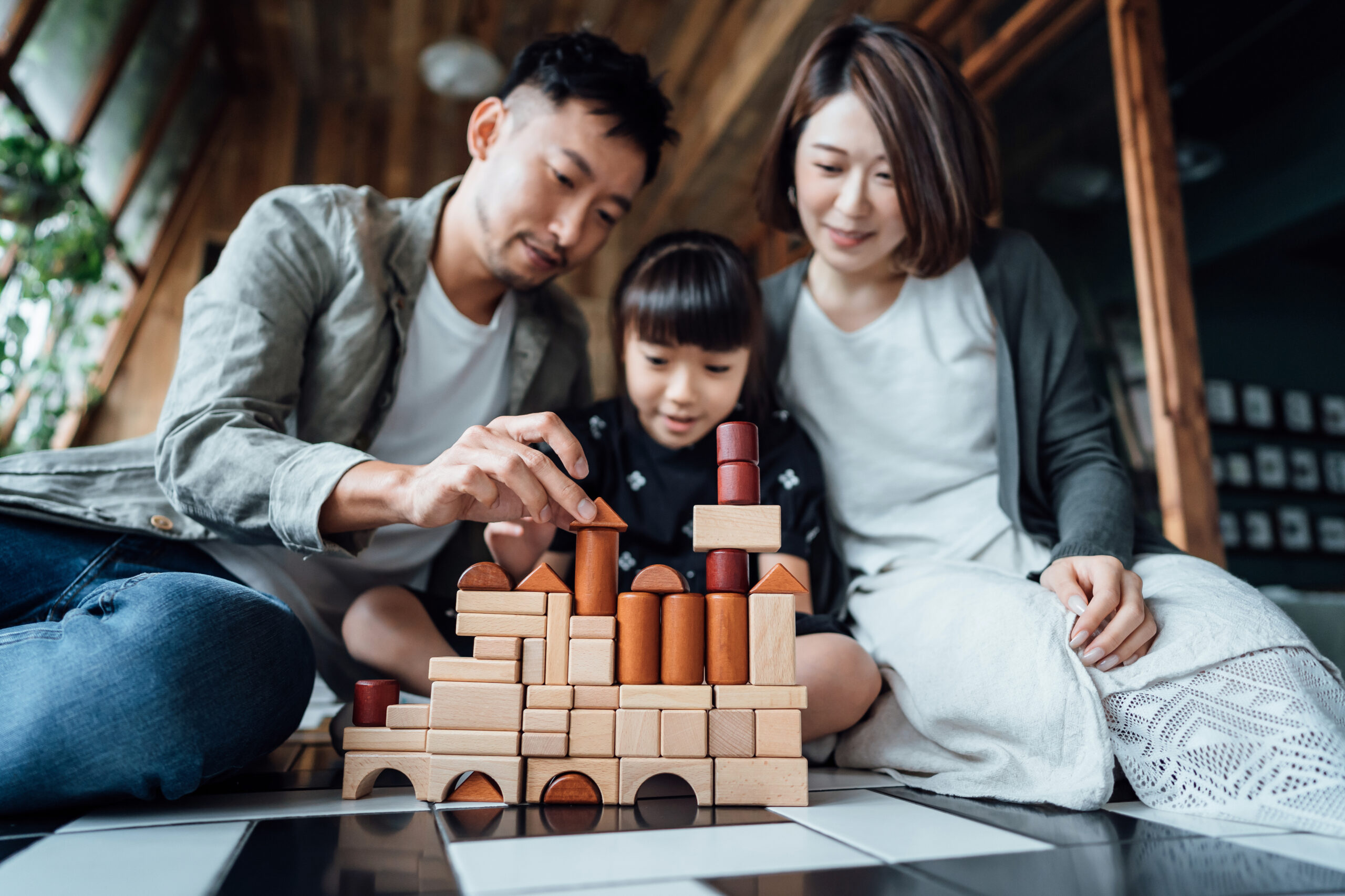 Joyful young Asian family sitting on the floor in the living room having fun playing wooden building blocks with daughter together at home